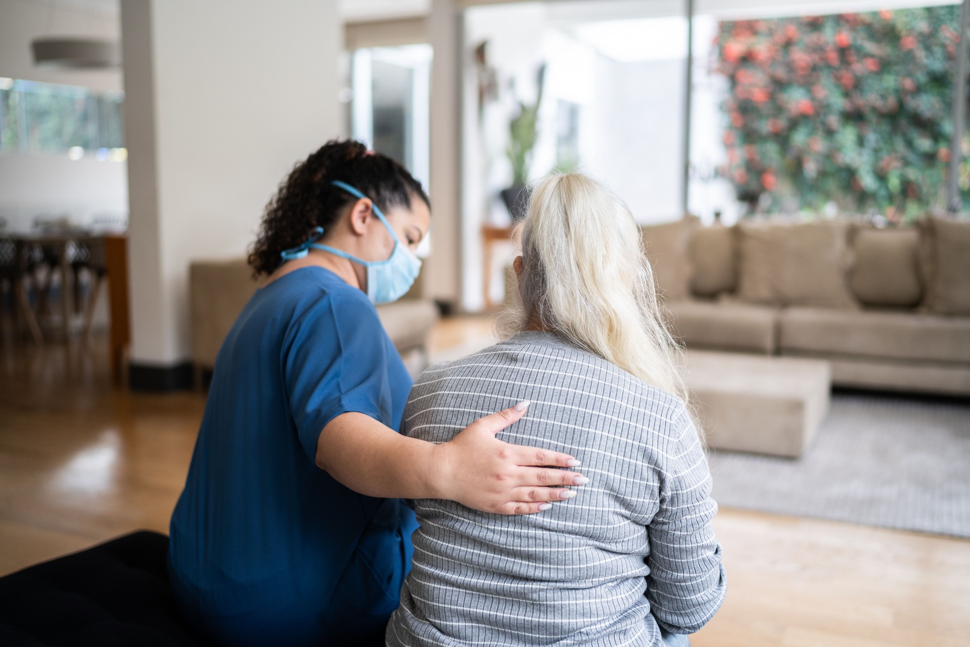 Home caregiver talking to and consoling senior patient at home - wearing protective face mask